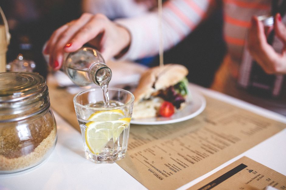 Close-up of water being poured into a glass with lemon on a restaurant table, next to a sandwich and sugar jar.