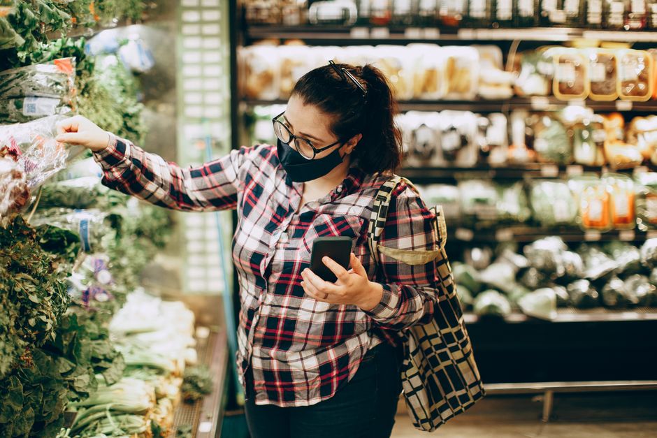 Woman wearing mask, shopping in grocery store, choosing fresh produce with smartphone in hand.