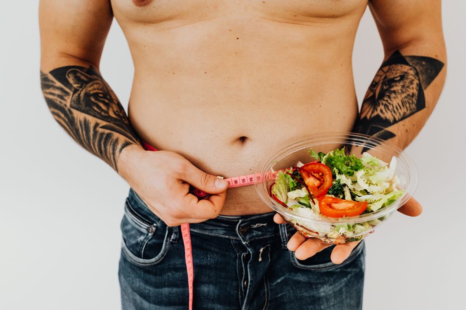 Tattooed man measuring waist while holding fresh salad, symbolizing healthy lifestyle and weight management.