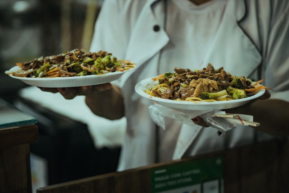 Chef presenting two plates of stir fry with noodles and vegetables in a restaurant kitchen.
