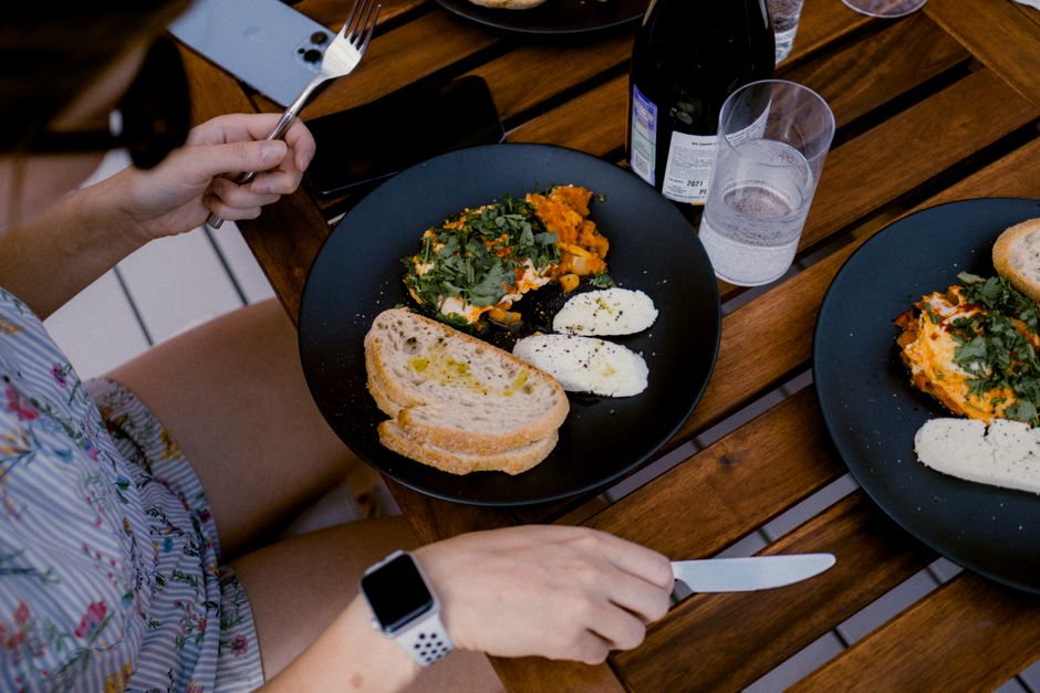 A casual outdoor brunch setting with fresh bread, cheese, and vegetables on dark plates.