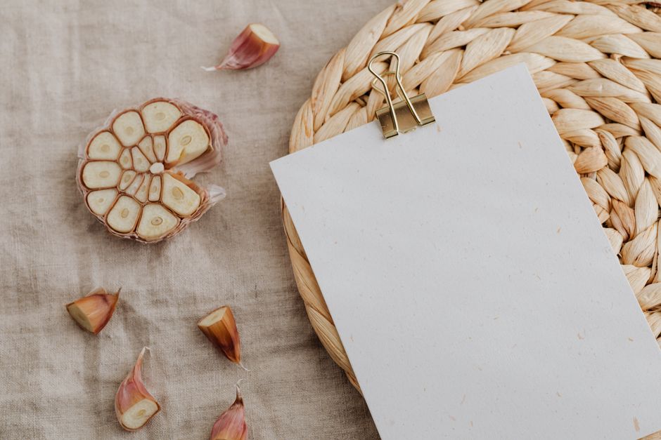 From above of sheet of white paper fixed with metal clip and put on wicker mat among cut raw head and cloves of garlic placed on linen towel