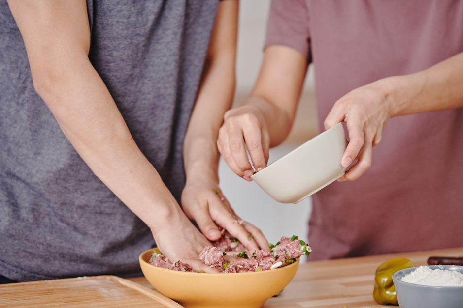 Two adults preparing a meat dish with fresh ingredients in a home kitchen.