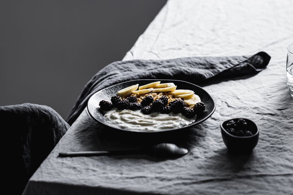 A tasteful black plate of yogurt with banana, blackberries, and granola on a table setting.